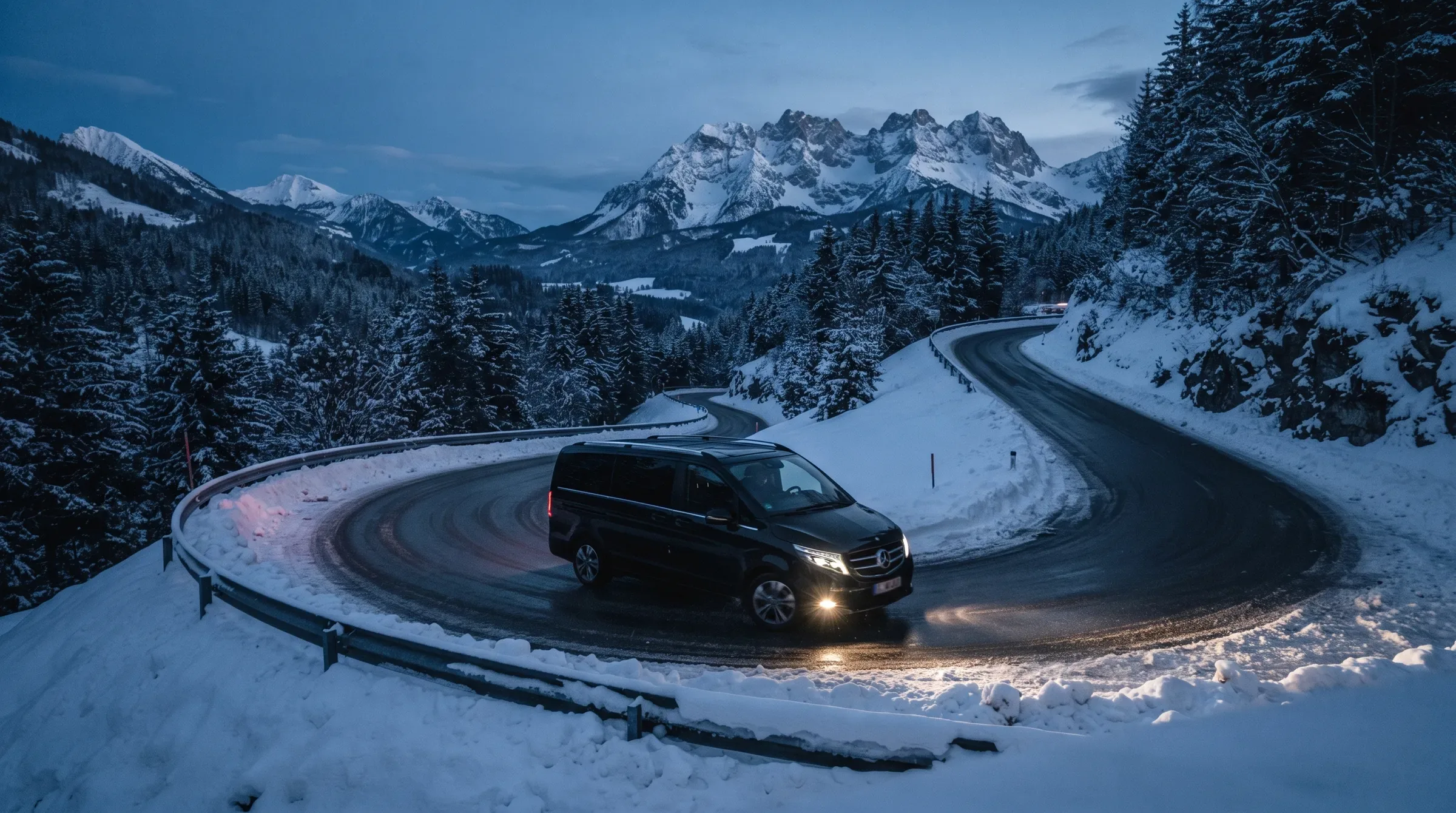 A Mercedes-Benz V-Class chauffeur vehicle navigating a winding Alpine mountain road at dusk, snow-capped peaks in the background.